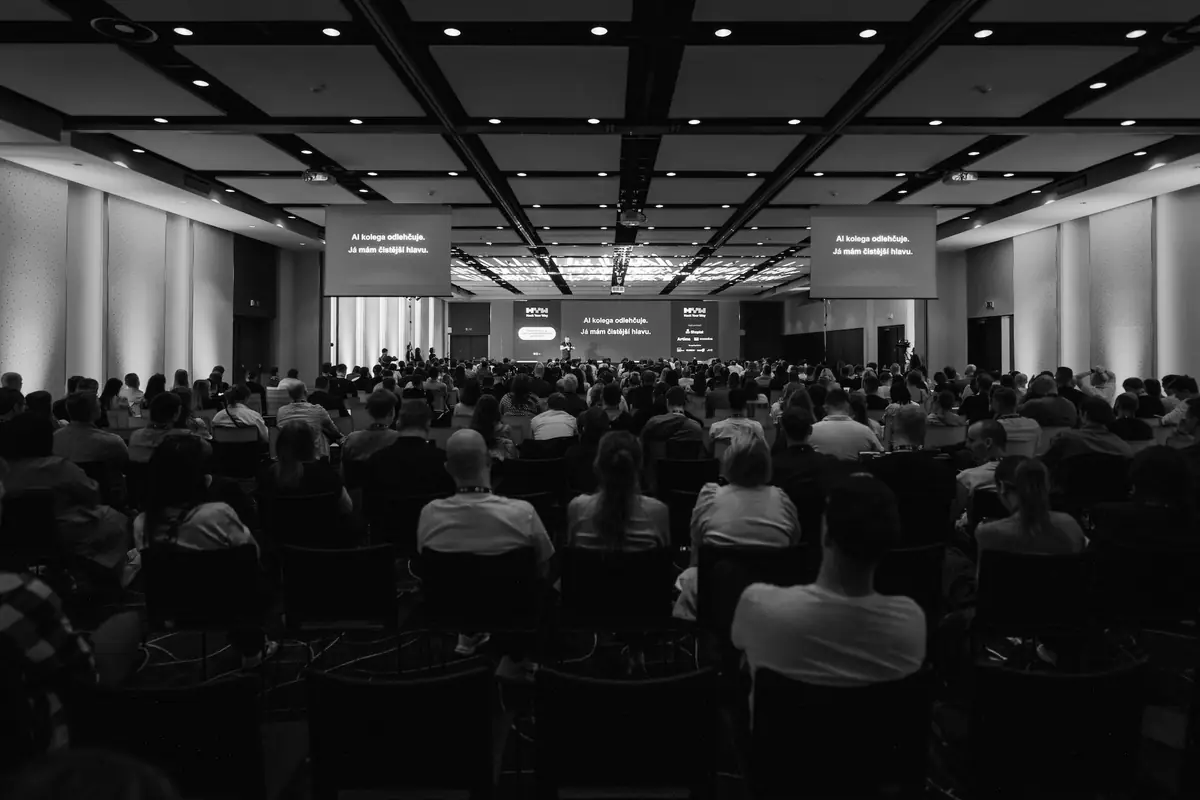 Packed hall at Hack Your Way 2026 in Brno, seen from the back rows; Jakub on stage under the slide 'AI kolega odlehčuje. Já mám čistější hlavu.'